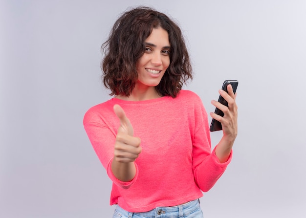 Souriante belle jeune femme tenant un téléphone mobile et montrant le pouce vers le haut sur un mur blanc isolé