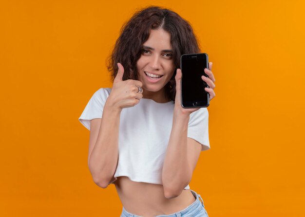 Souriante belle jeune femme tenant un téléphone mobile et montrant le pouce vers le haut sur un mur orange isolé avec espace copie