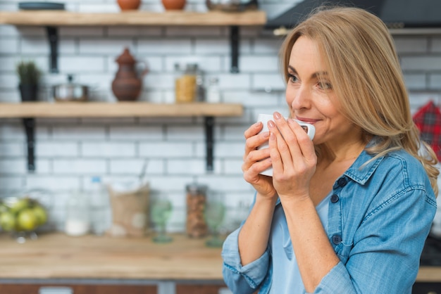 Souriante blonde jeune femme buvant du café de la tasse blanche dans la cuisine