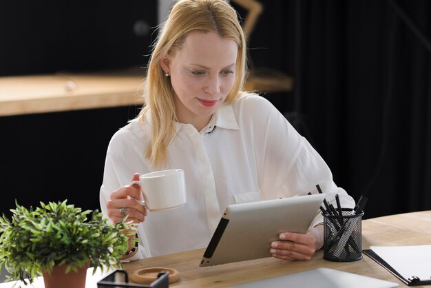 Souriante blonde jeune femme tenant une tasse de café en regardant une tablette numérique