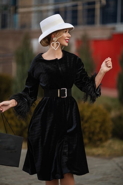 Photo gratuite souriante dame assez élégante en chapeau blanc et robe noire marchant dans la rue. concept de rue de la mode