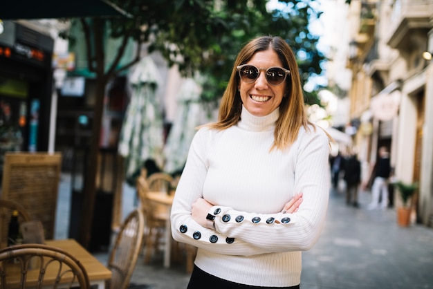 Souriante élégante jeune femme avec des lunettes de soleil près de café de rue dans la ville