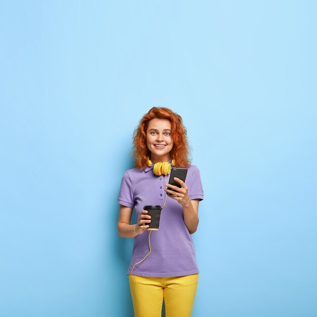 Souriante femme millénaire aux cheveux roux ondulés posant contre le mur bleu