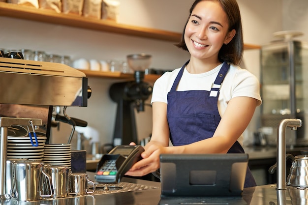 Souriante fille asiatique barista donnant au client pos terminal aider à payer avec une carte de crédit debout au comptoir
