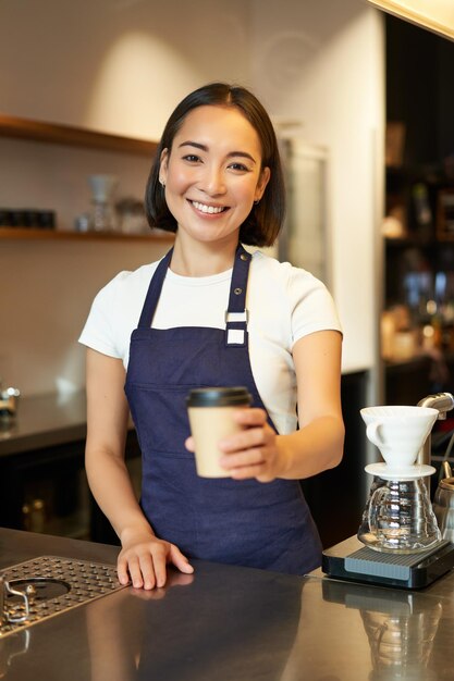 Souriante fille asiatique barista donnant l'ordre au client tenant une tasse de café à emporter portant un tablier de travail