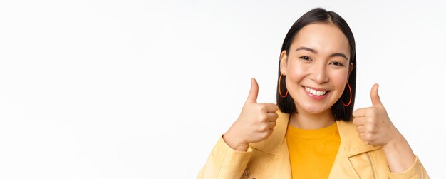 Souriante fille asiatique montrant les pouces vers le haut à la recherche d'approbation smth debout sur fond blanc