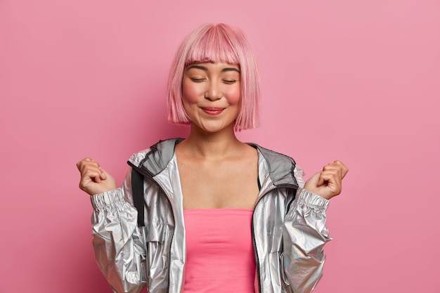 Souriante fille asiatique satisfaite aux cheveux rose bob, beauté naturelle, ferme les yeux, lève les mains les poings serrés, se sent très heureuse, vêtue d'un manteau d'argent à la mode,