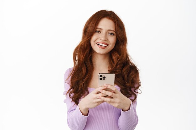 Souriante fille au gingembre bavardant, tenant un smartphone comme si elle faisait des achats en ligne, l'air heureuse tout en envoyant des messages, debout en blouse sur fond blanc