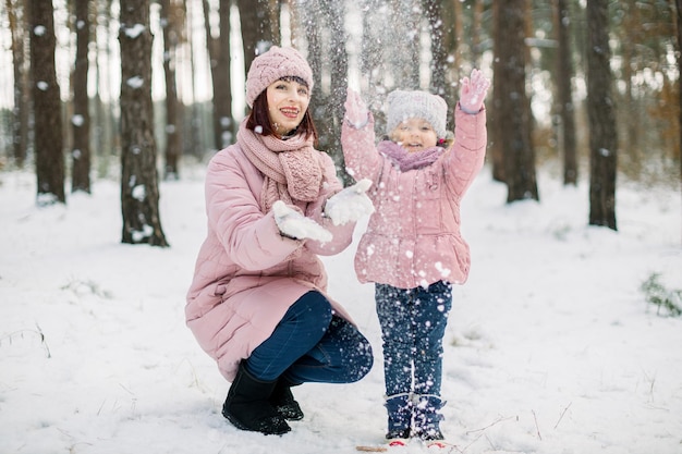 Souriante fille mignonne et heureuse qui jette de la neige et s'amuse lors d'une promenade avec sa jolie mère aimante. maman et enfant en manteaux roses s'amusant avec la neige dans la forêt d'hiver givrée.