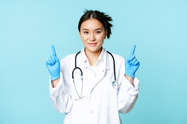 Souriante infirmière médecin asiatique dans des gants d'uniforme médical pointant les doigts vers le haut montrant les soins de santé i ...