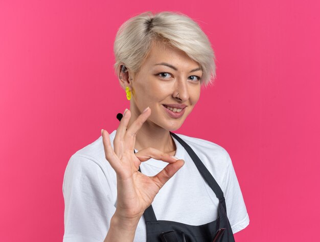 Souriante jeune belle femme barbier en uniforme montrant un geste correct isolé sur fond rose