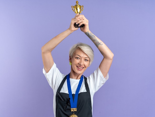 Photo gratuite souriante jeune belle femme barbier en uniforme portant la coupe du vainqueur de la médaille isolée sur fond bleu