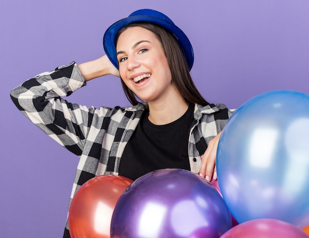 Souriante jeune belle femme portant un chapeau de fête debout derrière des ballons mettant la main sur la tête isolée sur le mur bleu