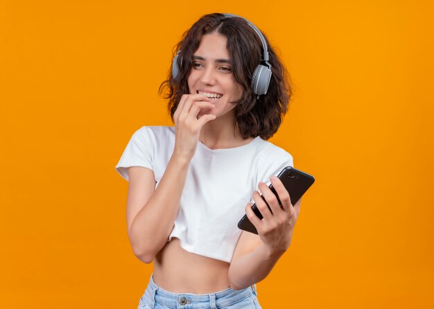 Souriante jeune belle femme portant des écouteurs tenant un téléphone mobile et mettant la main sur la lèvre sur un mur orange isolé avec espace copie