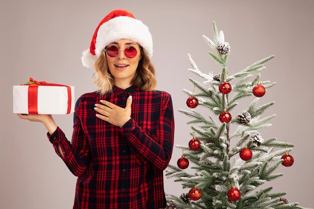 Souriante jeune belle fille debout à proximité de l'arbre de Noël portant un chapeau de Noël avec des lunettes tenant une boîte-cadeau mettant la main sur elle-même isolé sur fond blanc