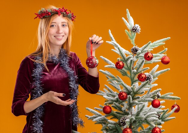 Souriante jeune belle fille debout à proximité de l'arbre de Noël portant une robe rouge et une couronne avec guirlande sur le cou tenant des boules de Noël isolées sur fond orange