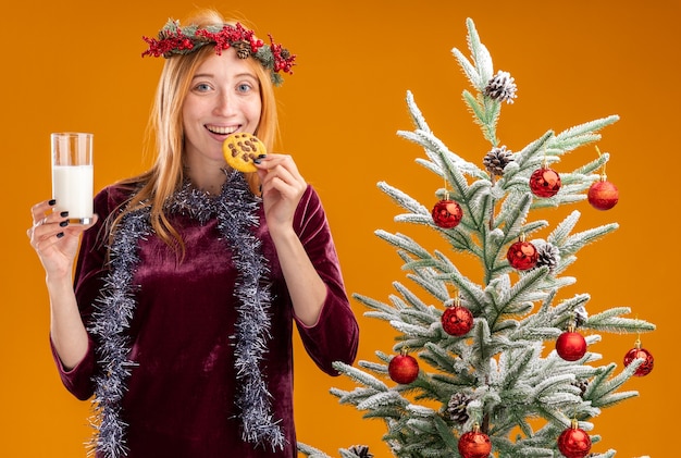 Souriante jeune belle fille debout à proximité de l'arbre de Noël portant une robe rouge et une couronne avec guirlande sur le cou tenant un verre de lait et essayer des cookies isolés sur fond orange