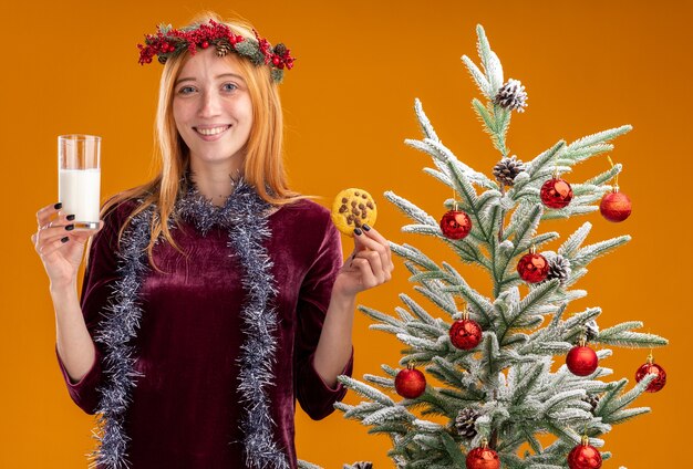 souriante jeune belle fille debout à proximité d'un arbre de noël vêtue d'une robe rouge et d'une couronne avec une guirlande sur le cou tenant un verre de lait avec des biscuits isolés sur un mur orange