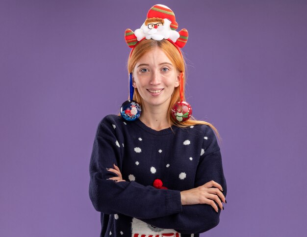 Souriante jeune belle fille portant chandail de Noël avec cerceau de cheveux de Noël accroché des boules de Noël sur l'oreille croisant les mains isolés sur fond violet