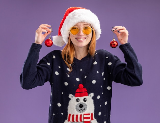 Souriante jeune belle fille portant chandail de Noël et chapeau avec des lunettes tenant des boules de Noël isolé sur fond violet