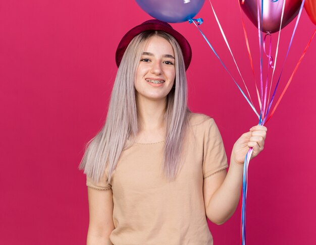 Souriante jeune belle fille portant un chapeau de fête avec des appareils dentaires tenant des ballons