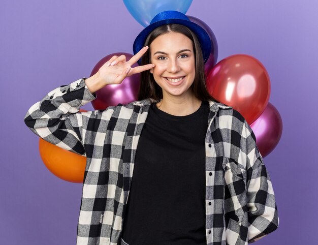 Souriante jeune belle fille portant un chapeau de fête debout devant des ballons montrant un geste de paix