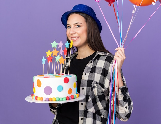 Souriante jeune belle fille portant un chapeau de fête tenant des ballons avec un gâteau isolé sur un mur bleu