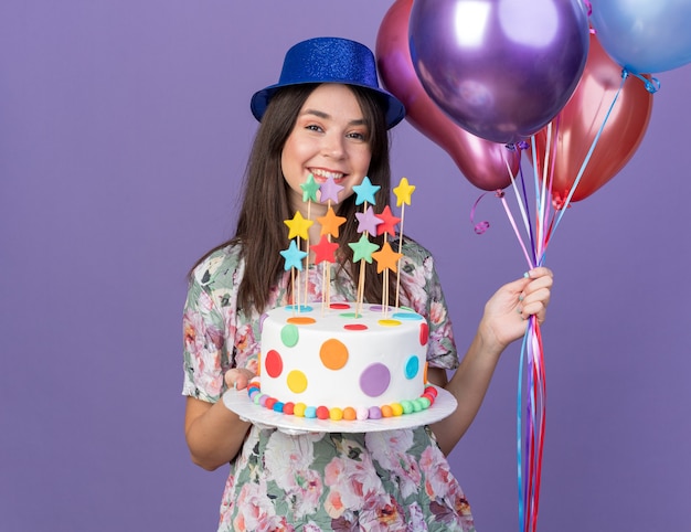 Souriante jeune belle fille portant un chapeau de fête tenant des ballons avec un gâteau isolé sur un mur bleu