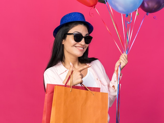 Souriante jeune belle fille portant un chapeau de fête tenant des ballons avec des points de sacs-cadeaux sur le côté