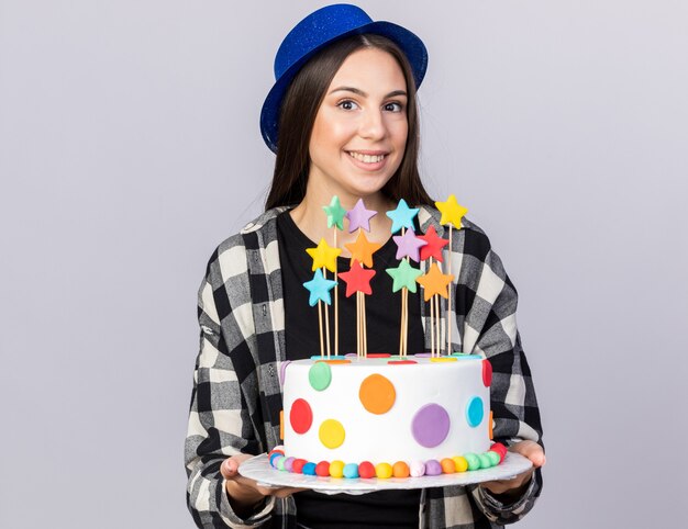 Souriante jeune belle fille portant un chapeau de fête tenant un gâteau isolé sur un mur blanc