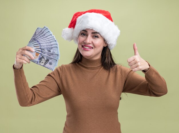 Souriante jeune belle fille portant un chapeau de Noël tenant de l'argent montrant le pouce vers le haut isolé sur fond vert olive