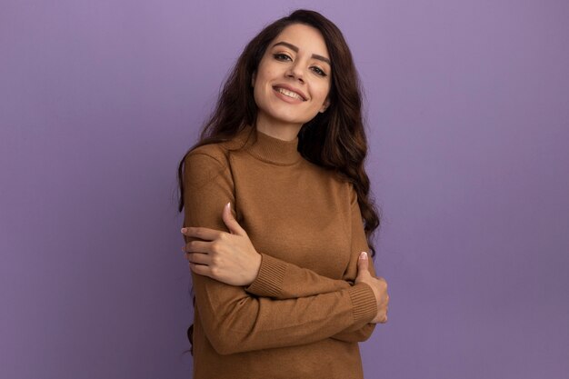 Souriante jeune belle fille portant un pull à col roulé marron croisant les mains isolés sur un mur violet avec espace copie