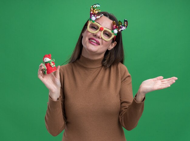 souriante jeune belle fille portant un pull marron et des lunettes de noël tenant un jouet de noël écartant la main isolée sur un mur vert