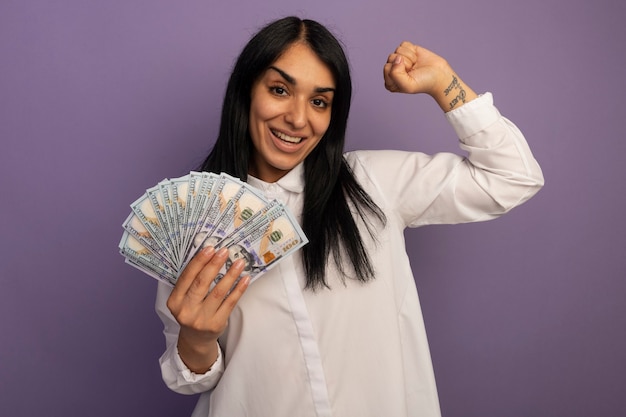 Souriante jeune belle fille portant un t-shirt blanc tenant de l'argent et montrant oui geste isolé sur violet