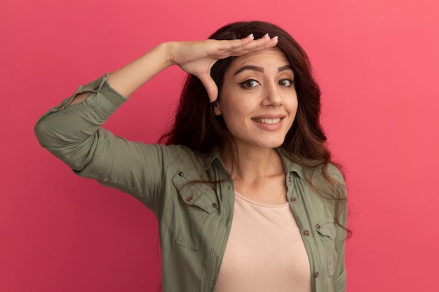 Souriante jeune belle fille portant un t-shirt vert olive montrant le geste de salut isolé sur un mur rose