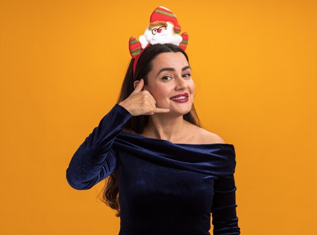 Souriante jeune belle fille vêtue d'une robe bleue et cerceau de cheveux de Noël tenant jouet montrant le geste d'appel téléphonique isolé sur fond orange