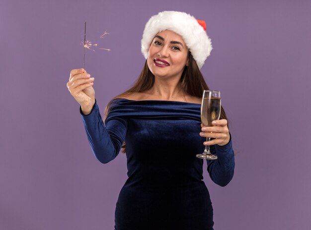 Souriante jeune belle fille vêtue d'une robe bleue et chapeau de Noël tenant des cierges magiques et tenant un verre de champagne à la caméra isolée sur fond violet