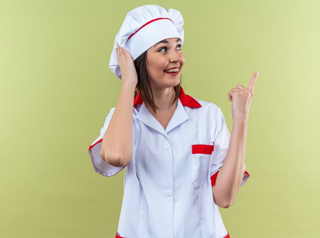 Souriante jeune cuisinière portant des points uniformes de chef sur le côté isolé sur un mur vert olive avec espace pour copie