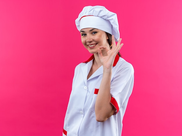 souriante jeune cuisinière portant un uniforme de chef montrant un geste correct isolé sur un mur rose