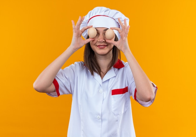 souriante jeune cuisinière portant l'uniforme du chef couvert les yeux avec des œufs isolés sur le mur orange