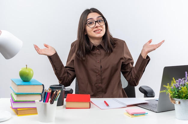 Souriante jeune écolière portant des lunettes est assise à table avec des outils scolaires écartant les mains