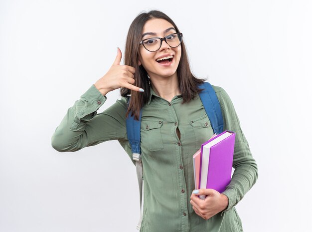 Souriante jeune écolière portant des lunettes avec un sac à dos tenant des livres montrant un geste d'appel téléphonique