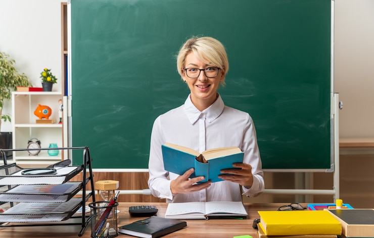 Photo gratuit souriante jeune enseignante blonde portant des lunettes assis au bureau avec des fournitures scolaires en classe tenant un livre ouvert regardant à l'avant