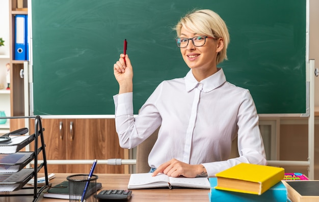 Photo gratuite souriante jeune enseignante blonde portant des lunettes assise au bureau avec des outils scolaires en classe pointant le doigt sur le bloc-notes regardant l'avant pointant vers le tableau avec un stylo