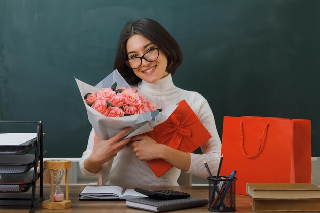 souriante jeune enseignante tenant des fleurs avec un cadeau assis au bureau avec des outils scolaires en classe