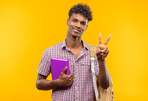 Souriante jeune étudiante afro-américaine avec sac à dos tenant un livre et gesticulant le signe de la victoire