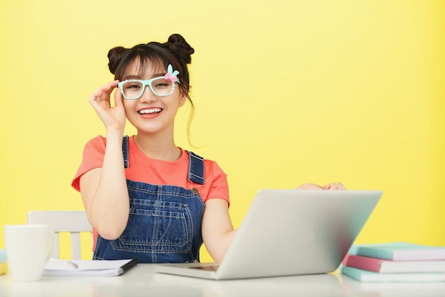 Souriante jeune étudiante asiatique dans des verres aux couleurs vives, assis au bureau avec ordinateur portable