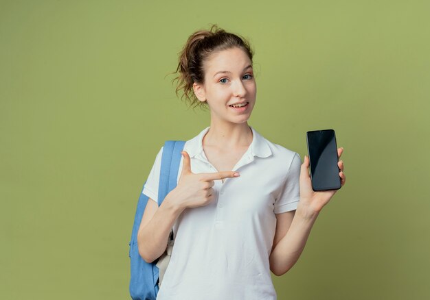 Souriante jeune étudiante jolie portant sac à dos montrant et pointant sur téléphone mobile isolé sur fond vert avec espace copie