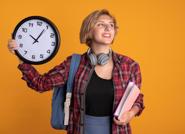 Souriante Jeune étudiante Slave Avec Des écouteurs Portant Un Sac à Dos Tient Un Livre D'horloge Et Un Ordinateur Portable Regardant De Côté