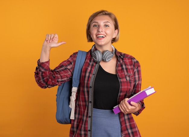 Souriante jeune étudiante slave avec des écouteurs portant un sac à dos tient un livre et un ordinateur portable sur elle-même